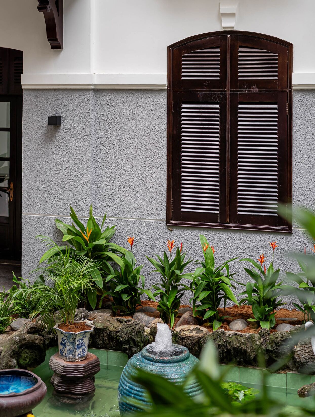 Edifici tradicional amb façana blanca i persianes de fusta fosca. Al davant, hi ha un jardí amb diverses plantes i una petita font.