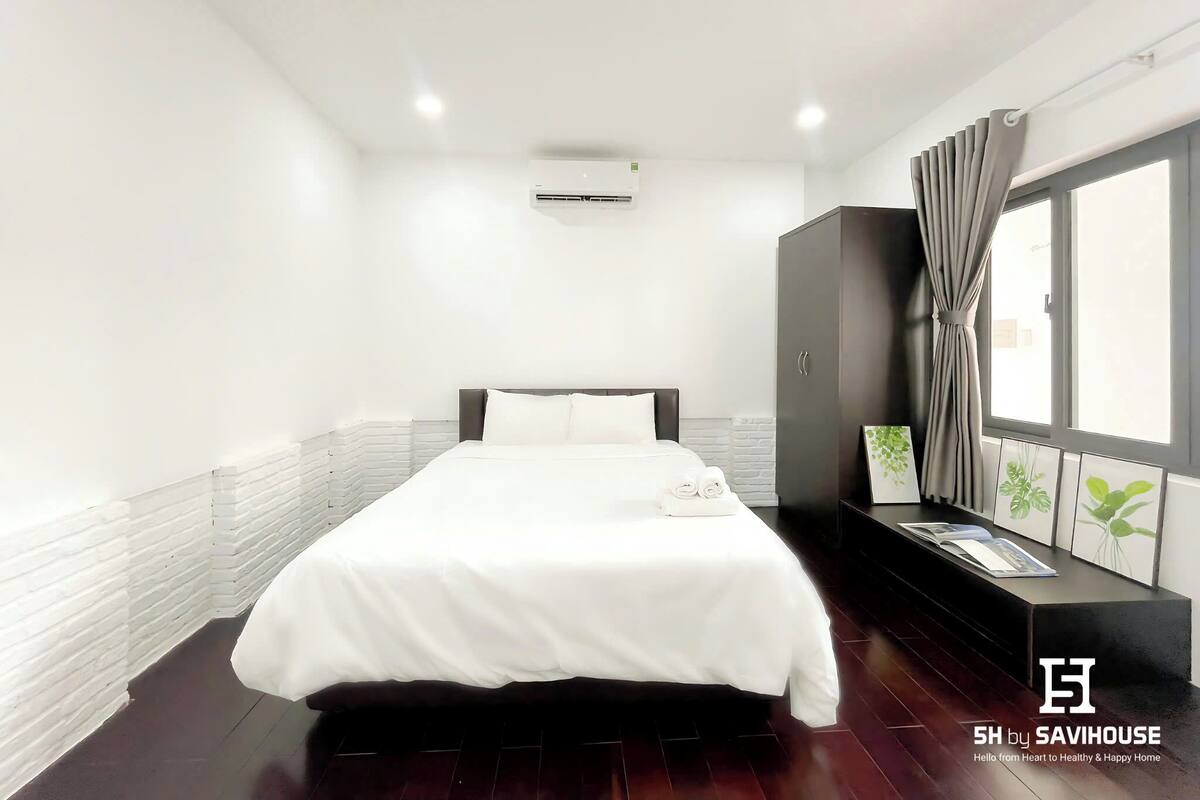 Une chambre d’hôtel avec un grand lit, une armoire, une fenêtre avec des rideaux et une petite table sur laquelle est posée une plante.