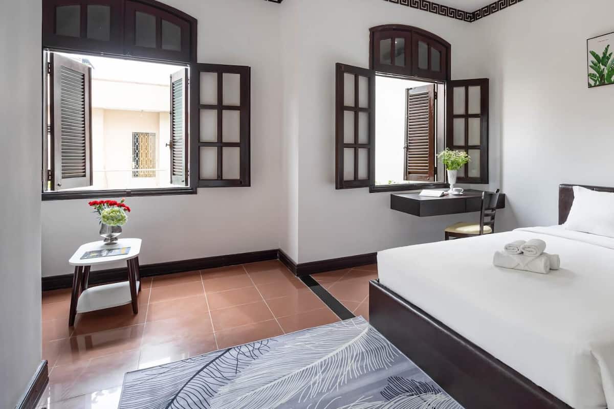 Une chambre d’hôtel avec un grand lit, un bureau avec une chaise, une petite table ornée d’un vase de fleurs et une fenêtre dont les volets sont ouverts.