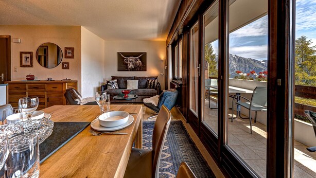 Un comedor con una mesa de madera dispuesta para una comida, un sofá al fondo y un gran ventanal con vistas a la montaña.
