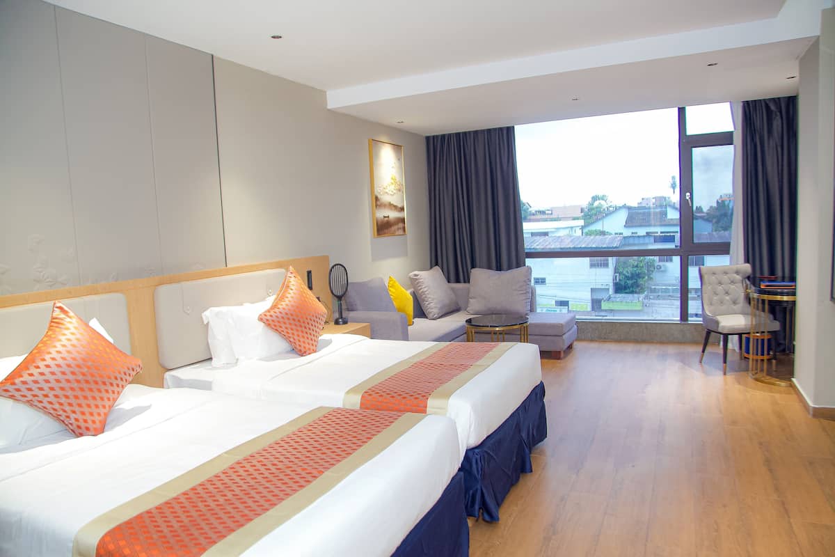 Habitación de hotel con dos camas, un sofá, una silla y un ventanal que ofrece vistas a edificios y zonas verdes.