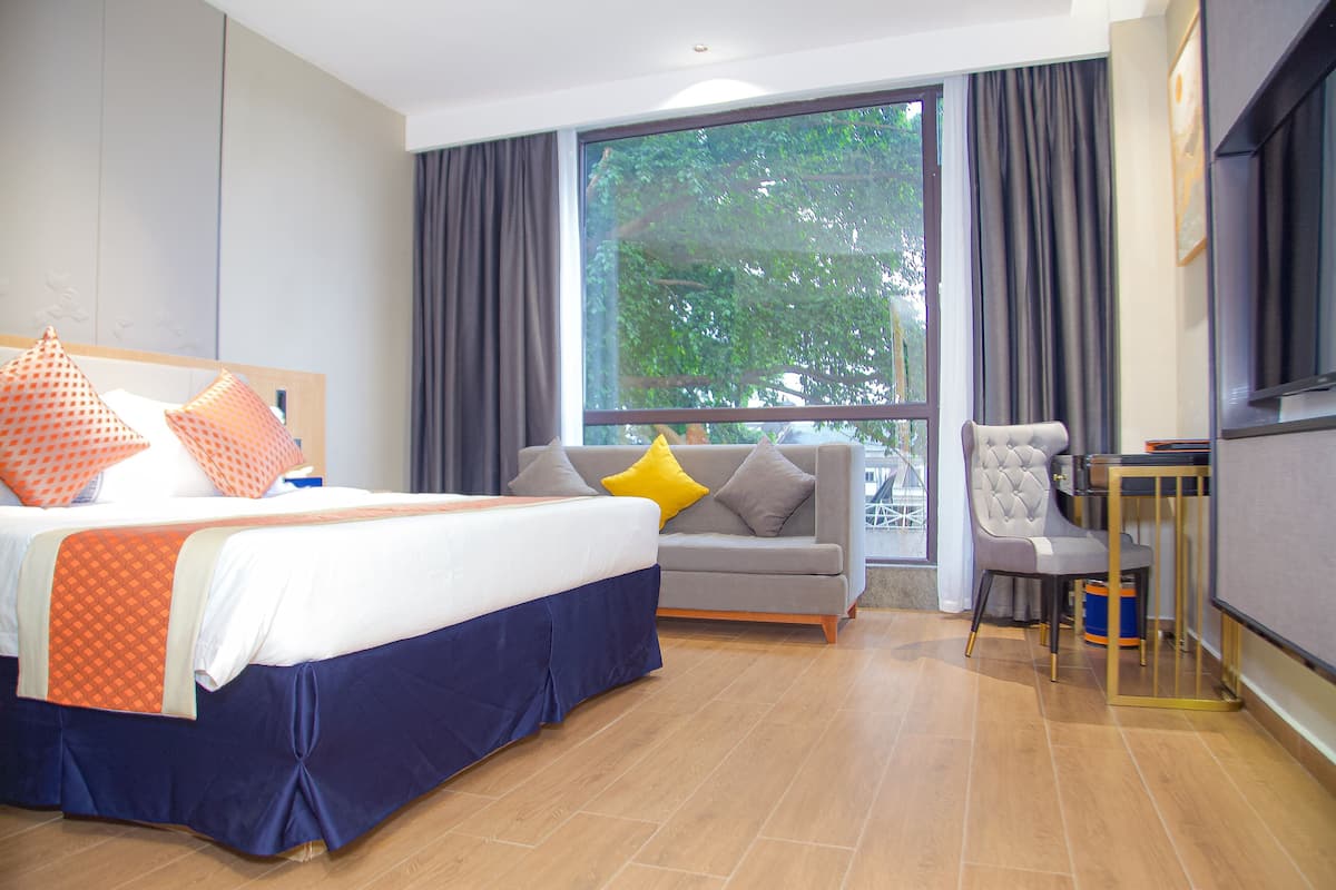 Una habitación de hotel con cama, sofá, sillón, televisor y un ventanal grande.