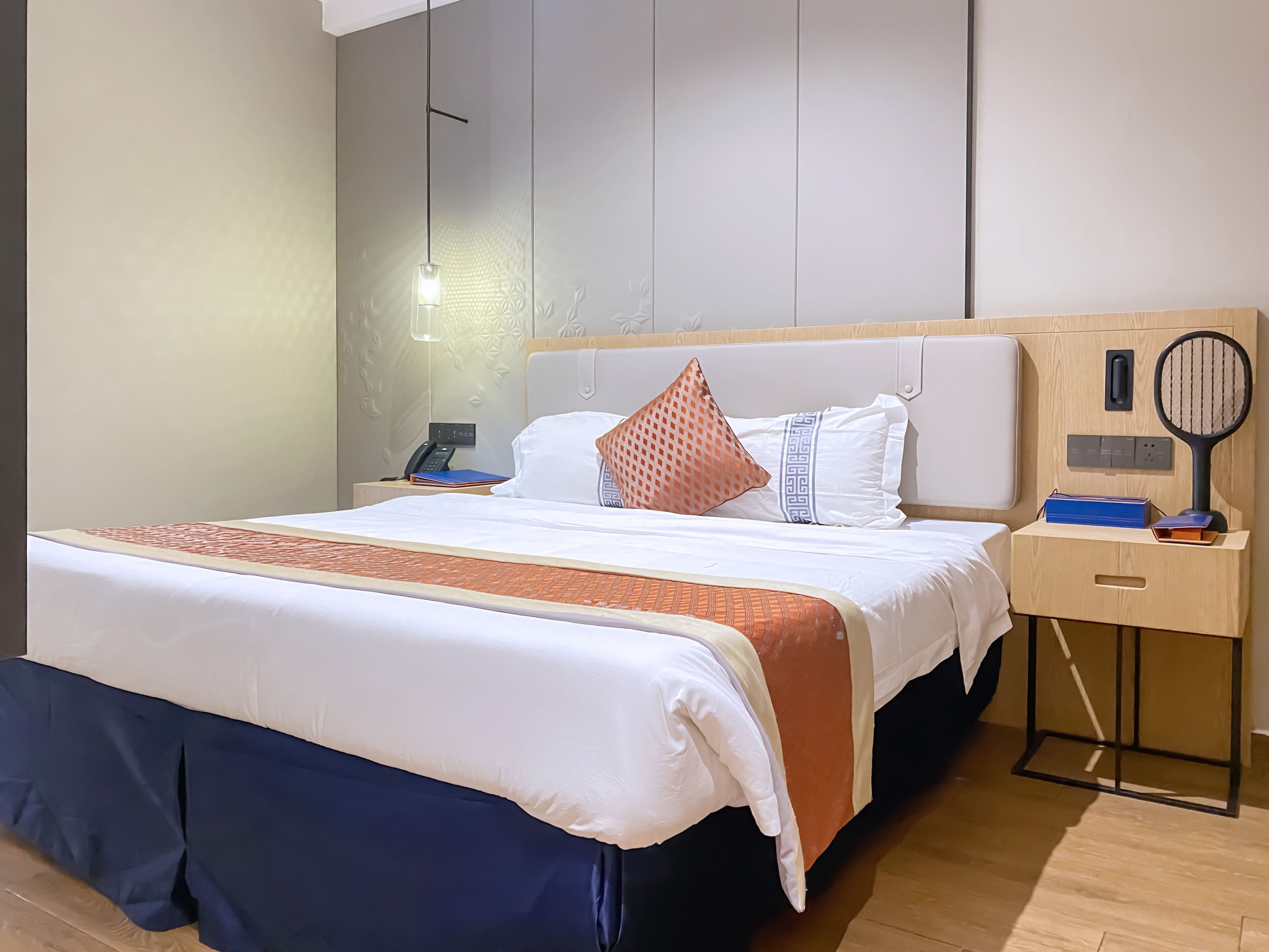 A neatly made bed with a white and orange bedspread, a wooden nightstand with a phone and a tennis racket, and a wall with a geometric pattern.