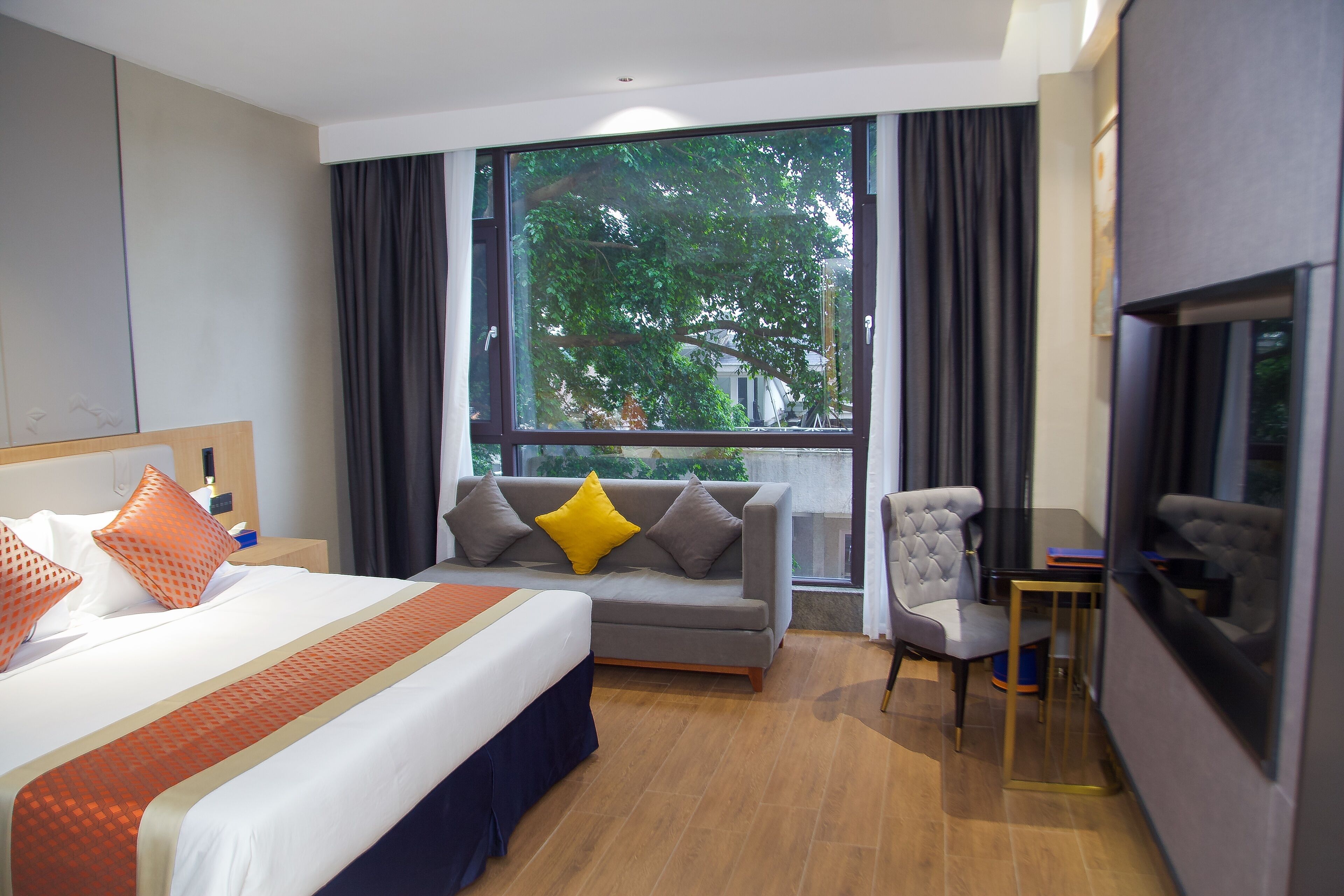 A hotel room with a bed, sofa, armchair, TV, and a large window with a view of trees.
