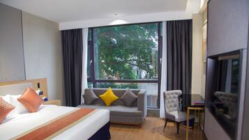 A hotel room with a bed, sofa, armchair, TV, and a large window with a view of trees.