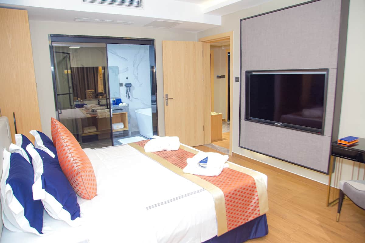 Habitación de hotel con cama, televisor, escritorio y baño visible a través de una puerta de cristal.