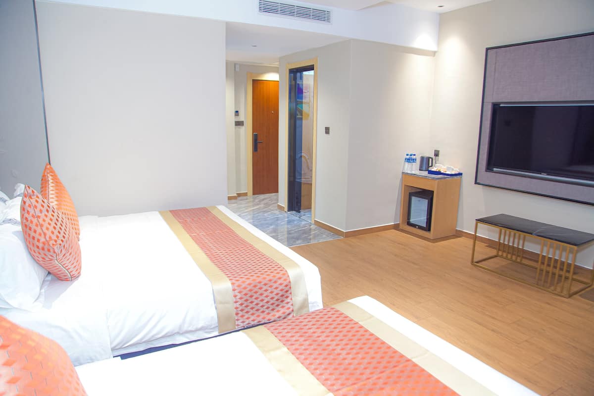 Habitación de hotel con una cama, un televisor, una mesita pequeña con una cafetera y una puerta que da a un pasillo.