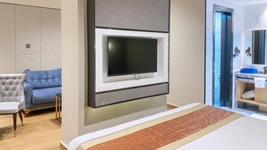 A modern hotel room with a large bed, a flat-screen TV mounted on the wall, a grey sofa, and a bathroom with a marble countertop. - FM HOTELS (Abidjan)