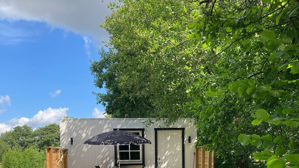 Eine kleine, weiße Holzkonstruktion mit Tür und Fenster, einem Tisch mit Stühlen und einem schwarzen Sonnenschirm auf der Veranda.