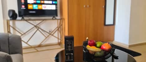 A living room with a TV on a stand, a coffee table with a fruit bowl, and a door in the background.