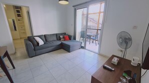 Un salon moderne avec un canapé d’angle gris, une table à manger, un ventilateur et une porte-fenêtre coulissante donnant sur une terrasse extérieure.