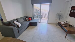 A living room with a grey sofa, a wooden side table, a fan, and a TV.