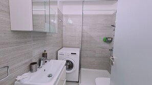 Moderni kylpyhuone, jossa on valkoinen pesuallas, pesukone, lasinen suihkuseinä ja wc.