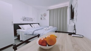 A hotel room with a bed, a table with fruit, a TV, and a window with curtains. - STAY Napa Living 41 (Ayia Napa)