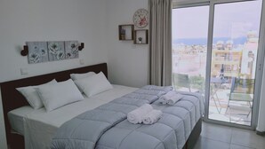 A bedroom with a bed, pillows, a quilted duvet, a clock, and a view of the city through a large window. - STAY Napa Living 39 (Ayia Napa)
