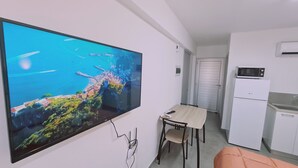 Una cocina moderna con microondas y refrigerador, un comedor con mesa y sillas, y un televisor grande montado en la pared que muestra una vista panorámica.