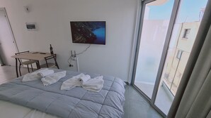 A hotel room with a bed, a desk, a chair, a painting, and a window with a view of buildings.