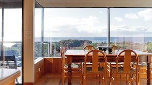 Ein Essbereich mit großem Tisch und Stühlen, Blick auf Meer und Hügel, und modernes Interieur.
