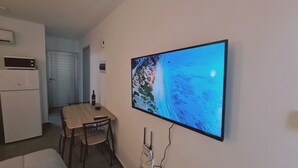 Une pièce dotée d’un grand téléviseur à écran plat fixé au mur, qui diffuse des images d’une plage, d’une table à manger en bois avec des chaises, d’un petit réfrigérateur et d’un four à micro-ondes.