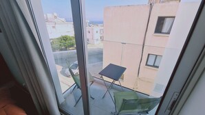 Un balcon avec une petite table et des chaises, donnant sur une rue bordée d’immeubles et offrant une vue sur un ciel dégagé.