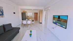 A modern living room with a sofa, a coffee table with plants, a dining area with chairs, a kitchen with cabinets, and a TV displaying a cartoon character. - STAY Azure Dream Apartment (Paralimni)