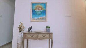 Ein Zimmer mit einer hellen Wand, einem gerahmten Gemälde einer Küstenlandschaft, einem kleinen Holztisch mit einer dekorativen Elefantfigur, einer Topfpflanze und einer Vase mit Trockenblumen.