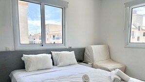 A bedroom with a bed, pillows, a bedside table, a chair, and two windows. - STAY Napa Aura Apartment (Ayia Napa)