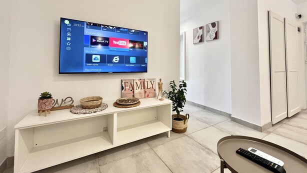 A modern living room with a mounted TV displaying a home screen, a white entertainment unit with decorative items, a potted plant, and a remote control on a tray. - STAY Napa Aura Apartment (Ayia Napa)