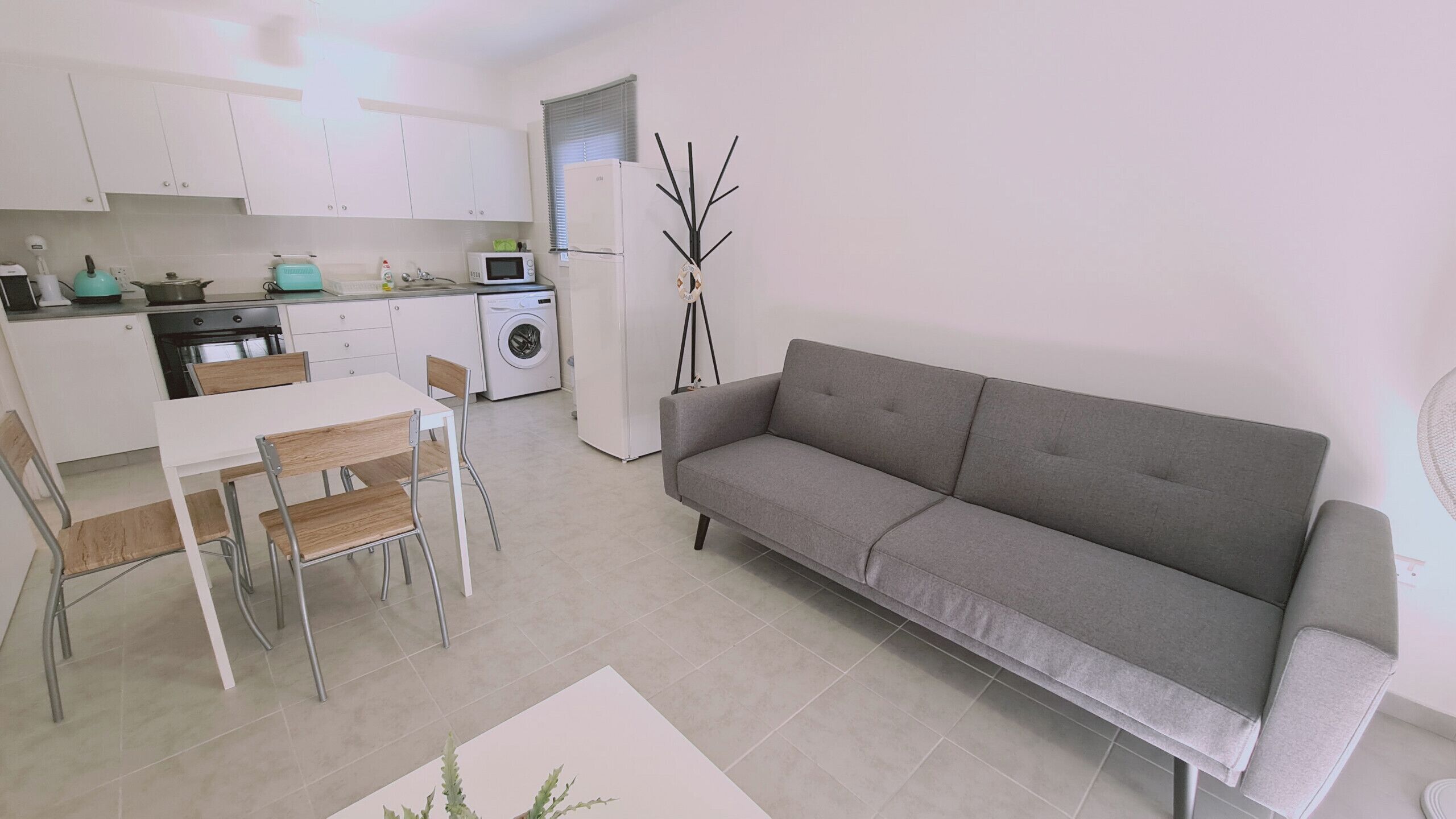 A modern kitchen with white cabinets, a dining table with chairs, a grey sofa, and a washing machine.