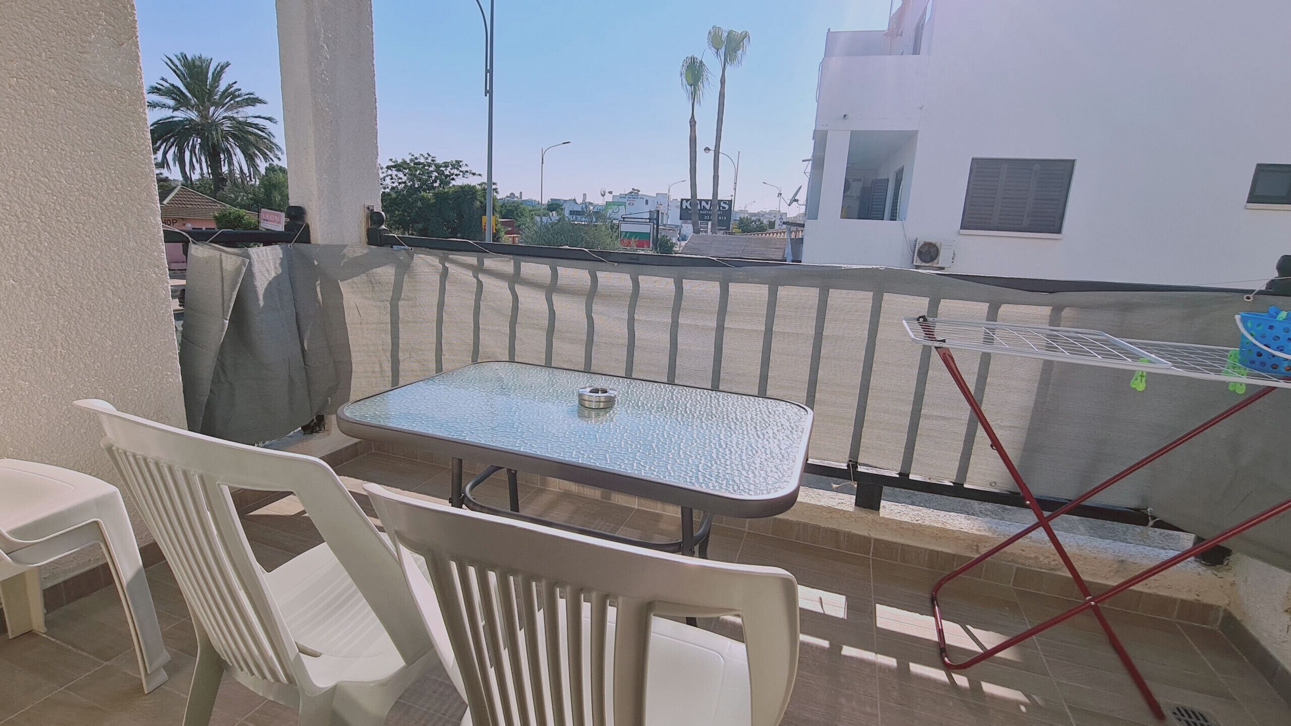 A balcony with a table and chairs, a drying rack, and a view of palm trees and buildings in the background.