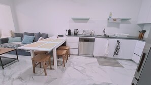 Una cocina compacta con lavavajillas, cafetera y pileta. Hay un comedor con mesa y sillas, y un sofá con almohadones.