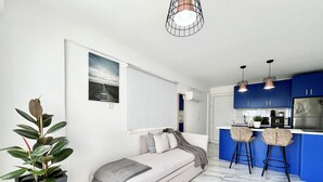 Een moderne woonkamer met een bank, een kamerplant en een keuken met blauwe kastjes en barstoelen.