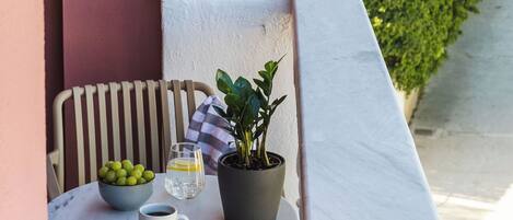 A balcony with a table, a chair, a potted plant, a bowl of fruit, and a cup of coffee.