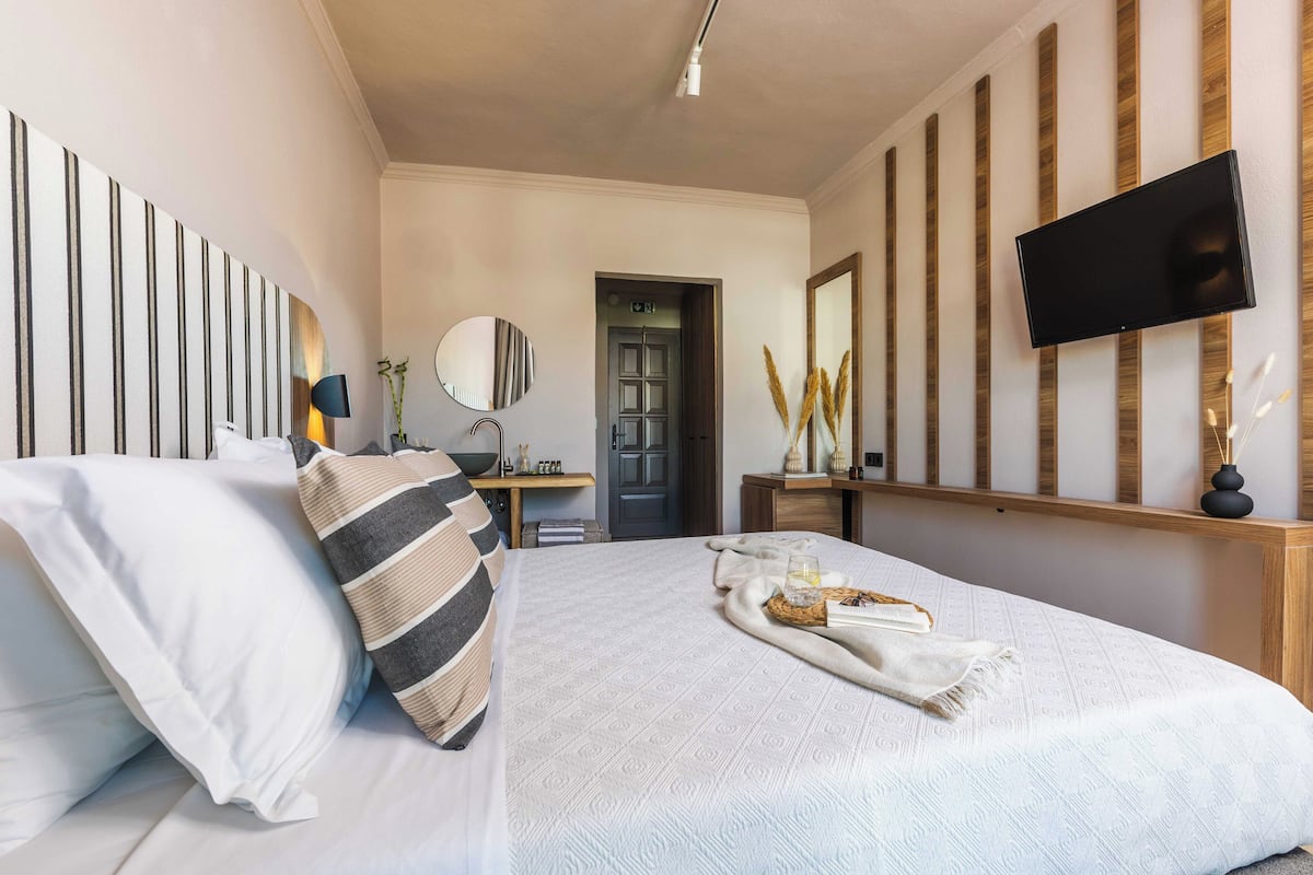 Une chambre à coucher comprenant un lit, un téléviseur à écran plat fixé au mur, une table de chevet avec une lampe, et une porte donnant sur une autre pièce.