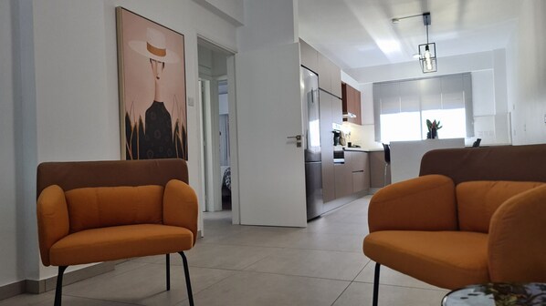 A modern interior with orange armchairs, a kitchen in the background, and a large painting on the wall.