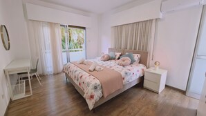 A bedroom with a bed, a desk, and a chair. There is a window with curtains, a bedside table with a lamp, and a wardrobe.