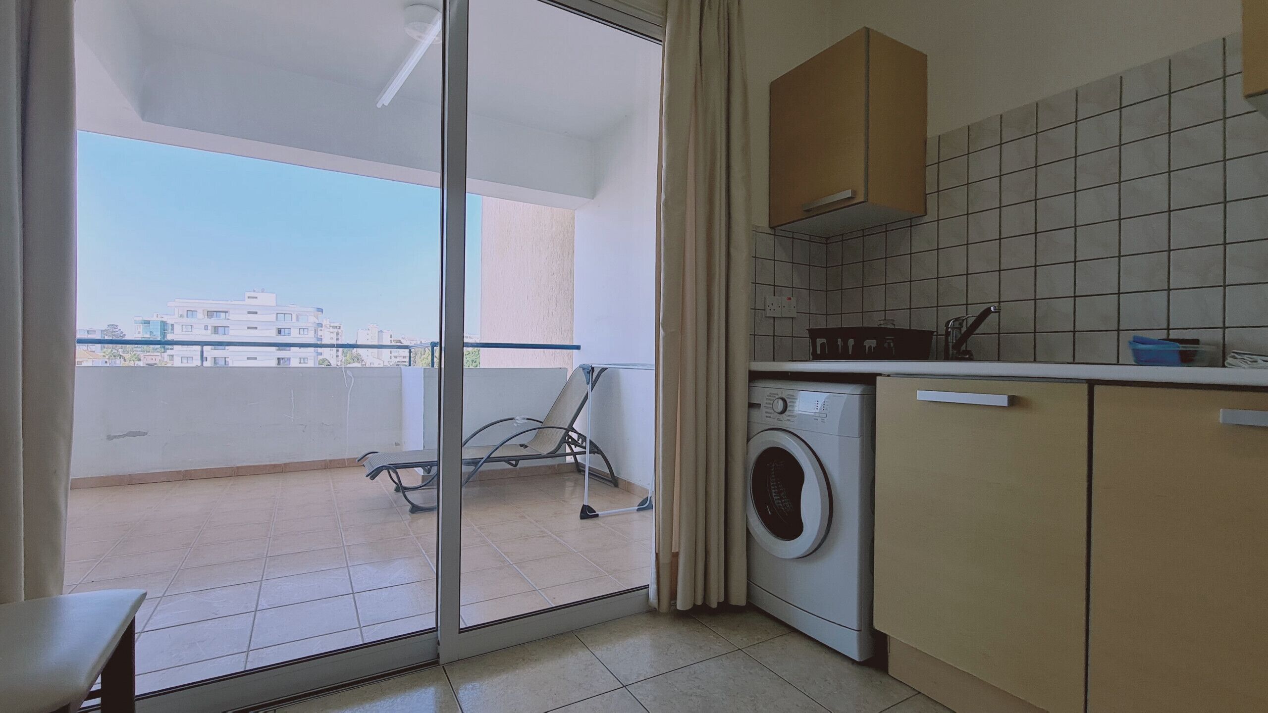 A kitchen with a washing machine, a chair, and a view of buildings through a glass door.