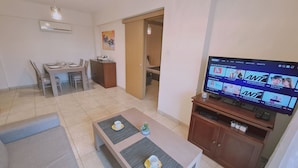 Una sala de estar con televisor mostrando un servicio de streaming, un comedor con mesa y sillas, y un sofá pequeño.