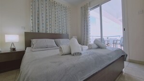 Habitación de hotel con una cama grande, una mesita de noche con lámpara y vistas al balcón.