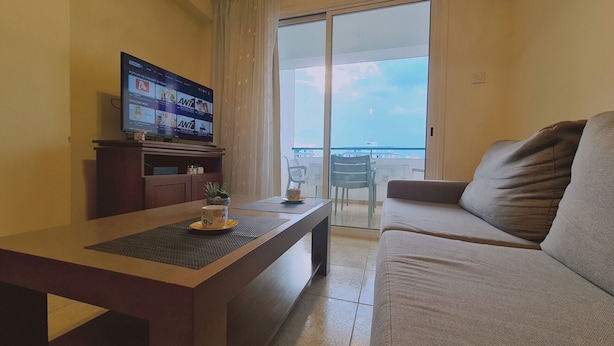 Una sala de estar con un sofá, una mesa de centro y un televisor mostrando un programa. Hay un balcón con vista al cielo y edificios.
