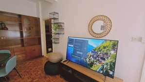 A living room with a television displaying a software update screen, a wooden cabinet, a wicker shelf with plants, and a round mirror on the wall. - STAY Antique Apartment (Larnaca)