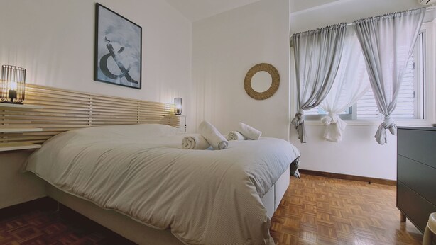 A bedroom with a bed, a bedside table, a lamp, a round mirror, and a window with curtains. - STAY Antique Apartment (Larnaca)