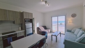 A compact kitchen with a microwave and refrigerator, a dining area with a table and chairs, a sofa, and a large window offering a view of the sea. - STAY Beach Wave Apartment (Larnaca)