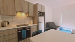 A modern kitchen with wooden cabinets, a built-in oven, a microwave, and a refrigerator. There is a table with chairs and a window with a view. - STAY Beach Wave Apartment (Larnaca)