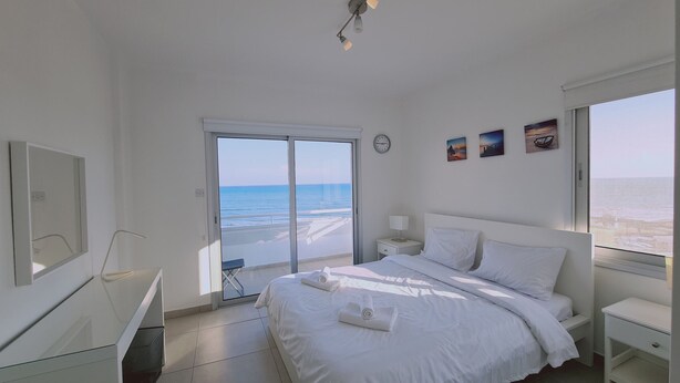A bedroom with a large bed, a nightstand, a desk, and a view of the ocean through a sliding door. - STAY Beach Wave Apartment (Larnaca)