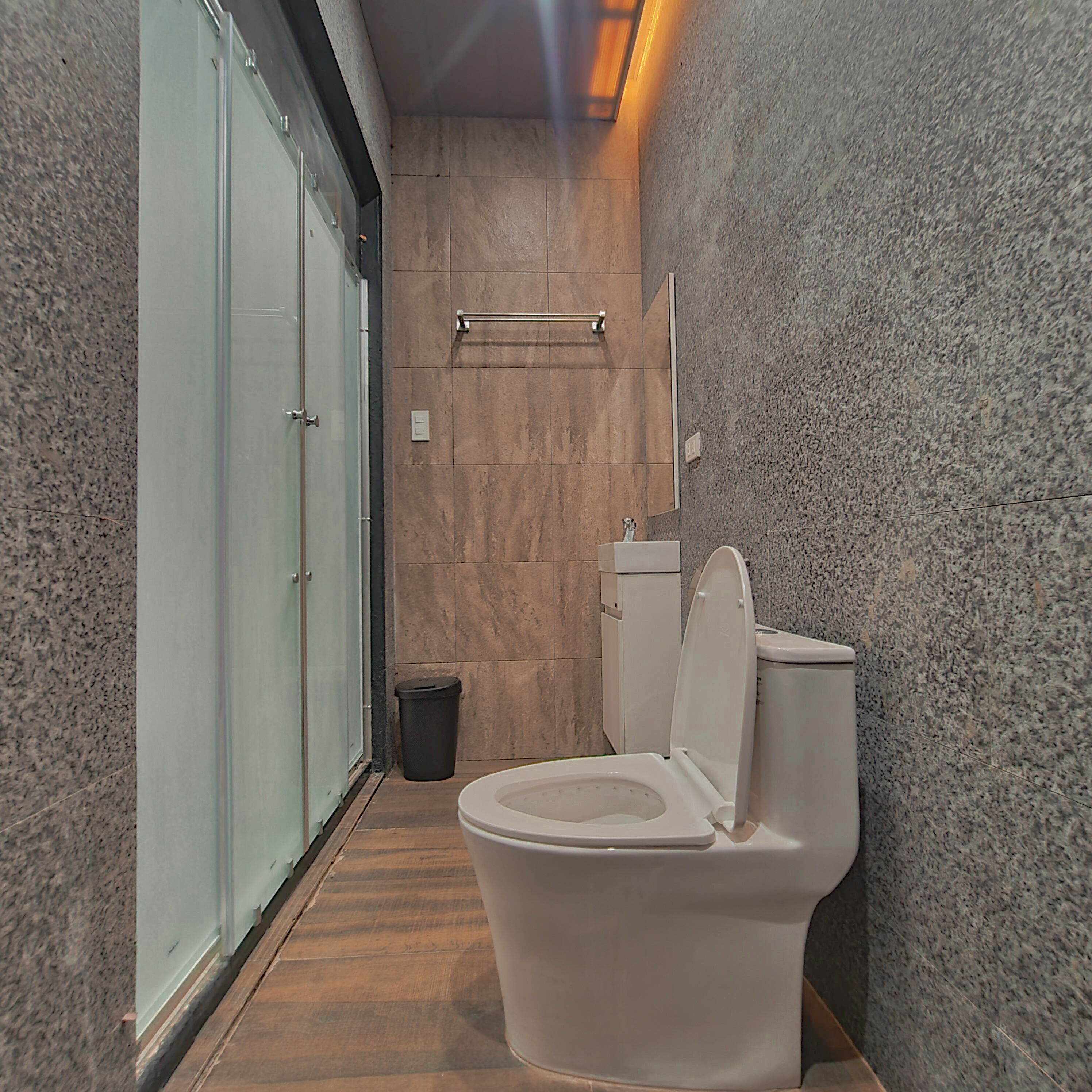 A narrow bathroom with a toilet, a trash bin, and a glass door leading to another room.