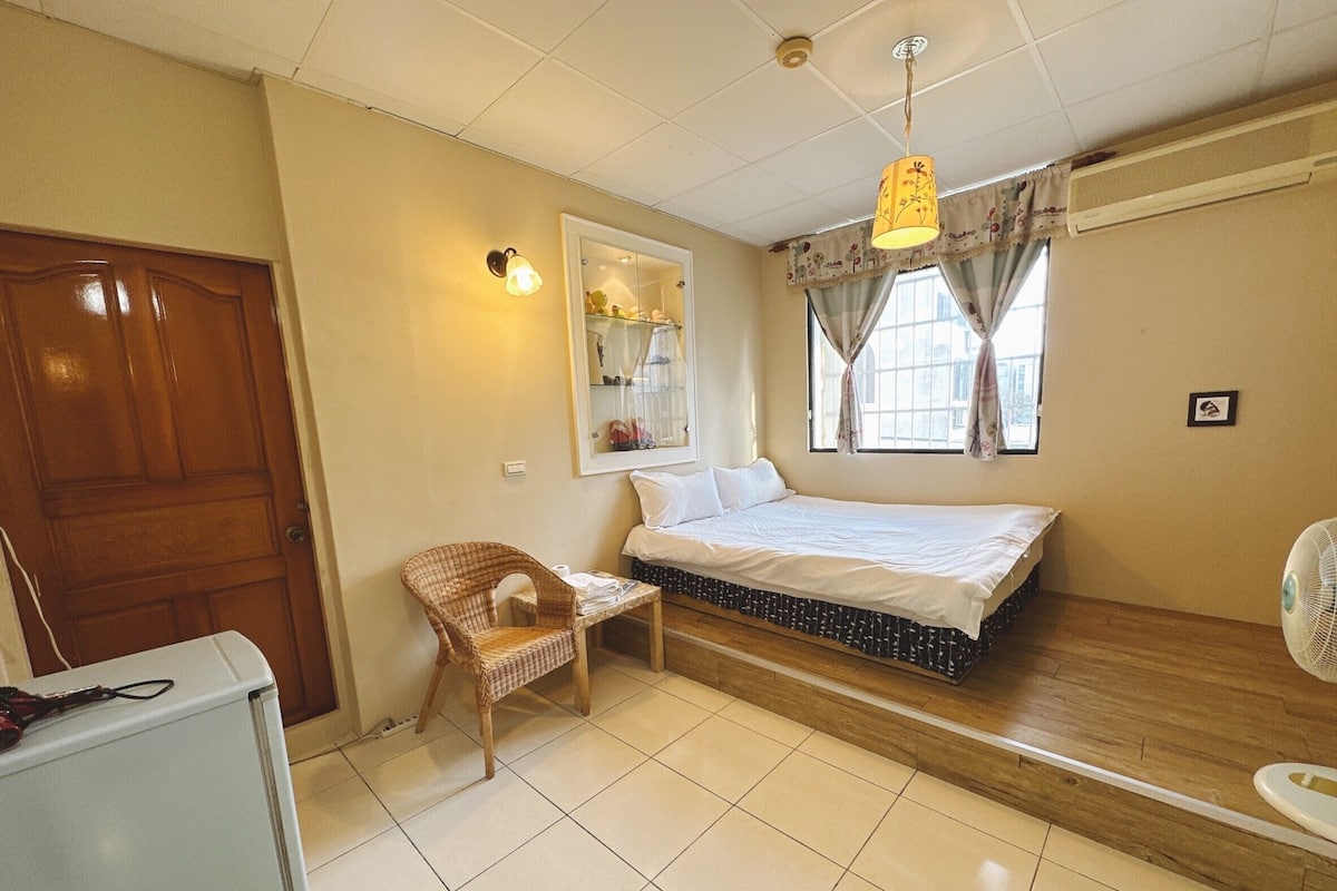 A compact bedroom with a bed, a chair, a small table, a mirror, and a window with curtains.