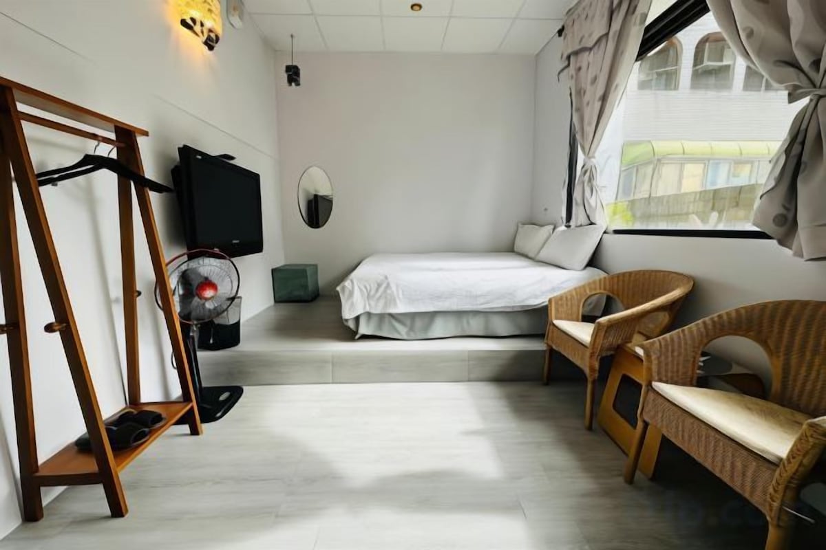 A bedroom with a bed, two wicker chairs, a standing fan, a TV, and a window with curtains.