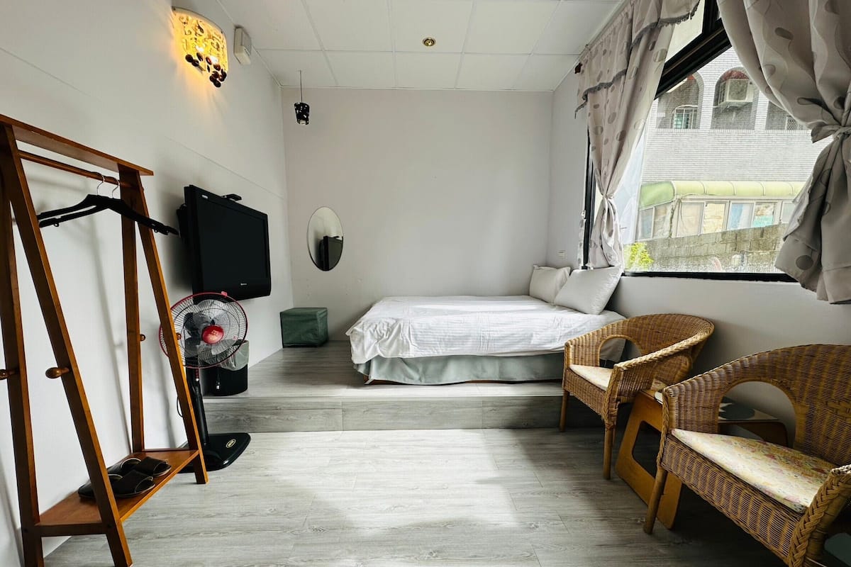 A bedroom with a bed, a fan, a chair, a TV, and a window with curtains.