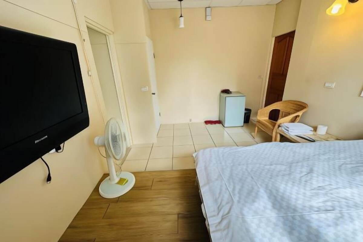 A hotel room with a bed, a television, a fan, a chair, and a small table.